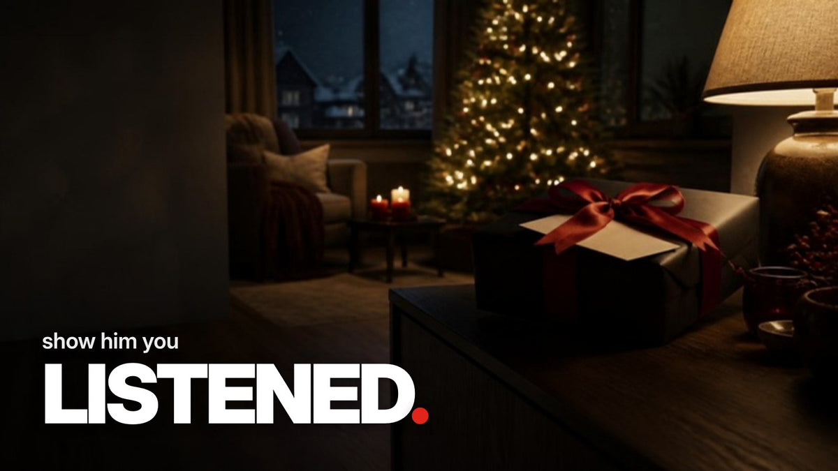 Schriftzug ‚show him you LISTENED.‘ über einem Foto eines in schwarzes Papier mit roter Schleife verpackten Geschenks auf einem Holzsideboard in einem abendlich beleuchteten Wohnzimmer mit Weihnachtsbaum.