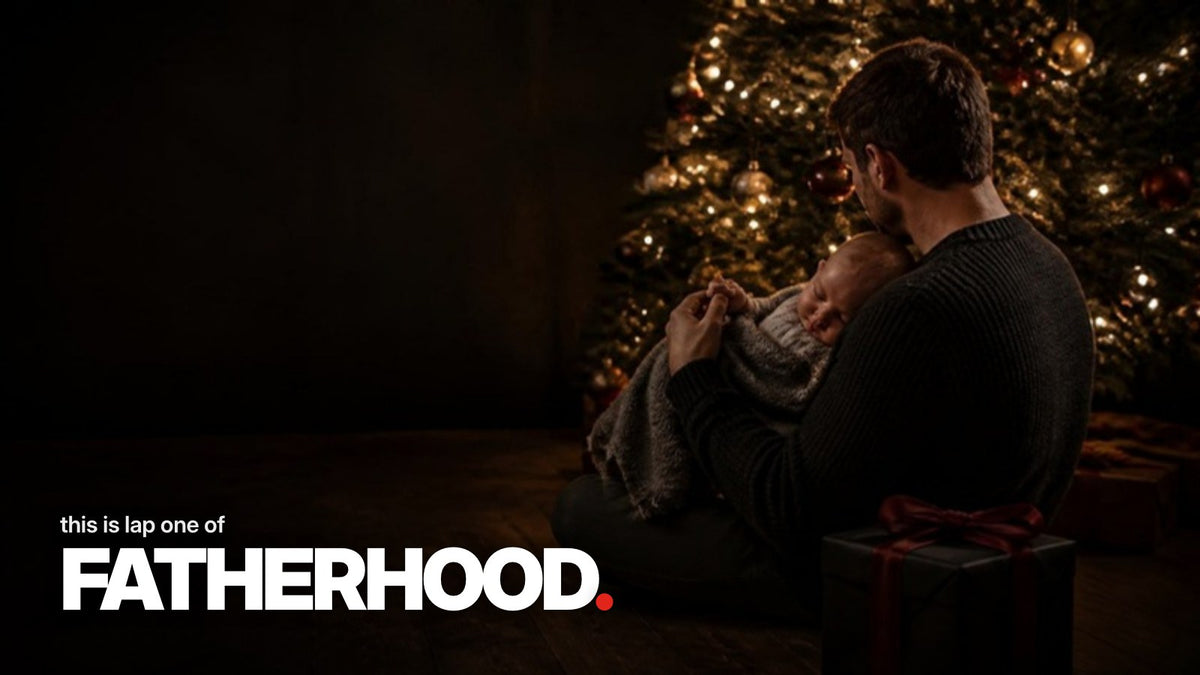 Scritta „this is lap one of FATHERHOOD.“ sopra una foto scura e suggestiva di un giovane padre visto di spalle che tiene un neonato addormentato sull’albero di Natale, accanto a un regalo incartato di nero con fiocco rosso.