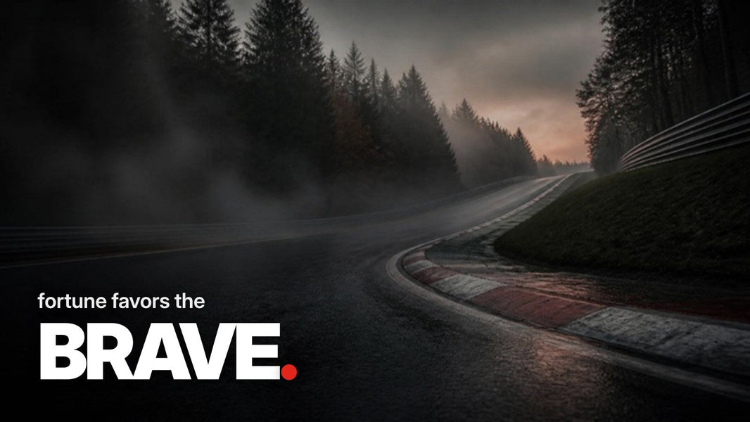 Text 'fortune favors the BRAVE.' over a photo of a wet, uphill racing curve with red-and-white curbs in a misty coniferous forest at dusk.