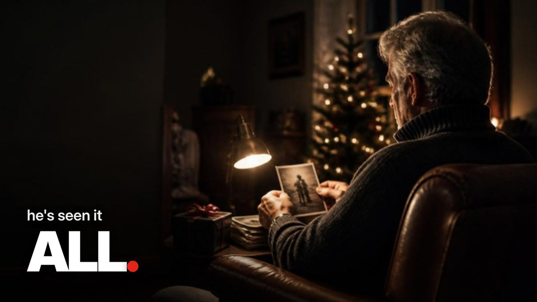 Schriftzug ‚he's seen it ALL.‘ über einem Foto eines älteren Mannes, der an einem Dezemberabend im warm beleuchteten Wohnzimmer im Ledersessel ein altes Foto betrachtet, neben ihm ein schwarz verpacktes Geschenk mit roter Schleife.
