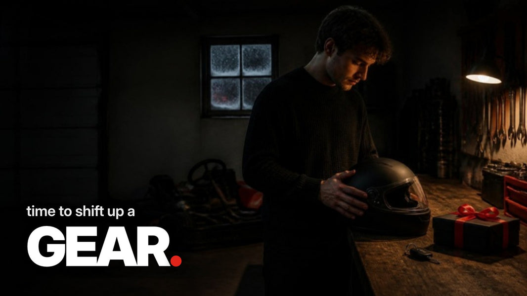 Schriftzug ‚time to shift up a GEAR.‘ über einem dunklen Foto einer Garage im Dezember mit einem schwarzen Helm, einem in schwarzes Papier mit roter Schleife verpackten Geschenk und einem Autoschlüssel auf einer Werkbank.