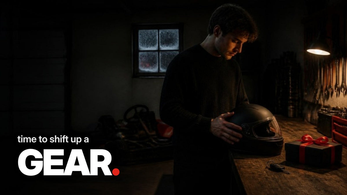 Schriftzug ‚time to shift up a GEAR.‘ über einem dunklen Foto einer Garage im Dezember mit einem schwarzen Helm, einem in schwarzes Papier mit roter Schleife verpackten Geschenk und einem Autoschlüssel auf einer Werkbank.