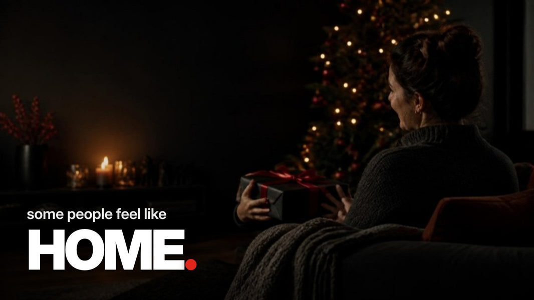Tekst ‚some people feel like HOME.‘ boven een foto waarop kinderhanden een vrouw in een donkere, kaarsverlichte woonkamer met Kerst een zwart verpakt cadeau met rode strik aanbieden.