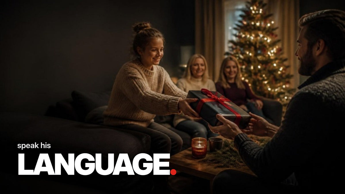 Schriftzug ‚speak his LANGUAGE.‘ über einem Foto, auf dem beim Familienwichteln im abendlichen Wohnzimmer ein Geschenk in schwarzem Papier mit roter Schleife überreicht wird, im Hintergrund ein beleuchteter Weihnachtsbaum.